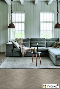 Moduleo Laurel Oak 51937 flooring in a traditional style living room with a low backed grey corner sofa and small round wooden table.