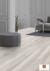 Light coloured V4 Fenland Oak laminate flooring in a modern living room with a grey low backed sofa and matching round foot stool.