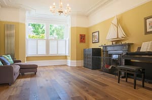 Simply Bespoke Collection flooring in a traditional style living space with a low level grey sofa and upright piano next to a wood burning fireplace.