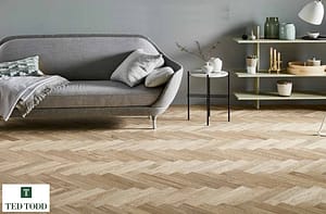 Tan Coloured Ted Todd Herringbone Sugar Cane European Oak flooring in a contemporary living room with a low level grey sofa and metal framed side table.