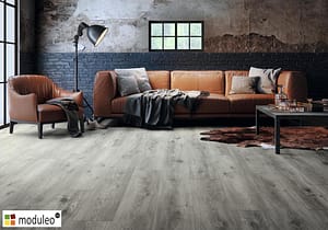 Moduleo Sierra Oak 58936 flooring in a warehouse style living space with a low backed tan leather sofa and matching chair.
