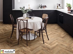 Moduleo Country Oak 54852 flooring in a contemporary style kitchen with black fronted units and a round wooden table and chairs.